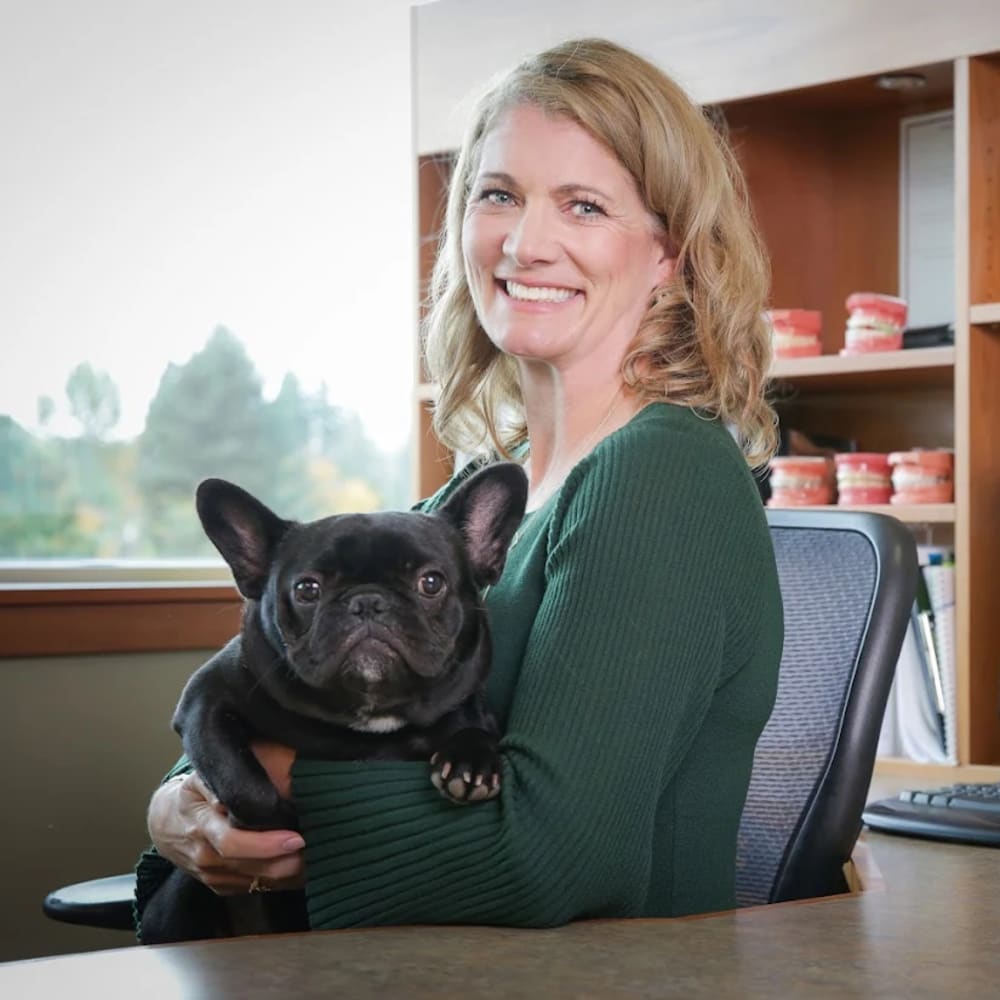 Dr. Lyons holding a black French Bulldog while sitting at an office desk and smiling at the camera.