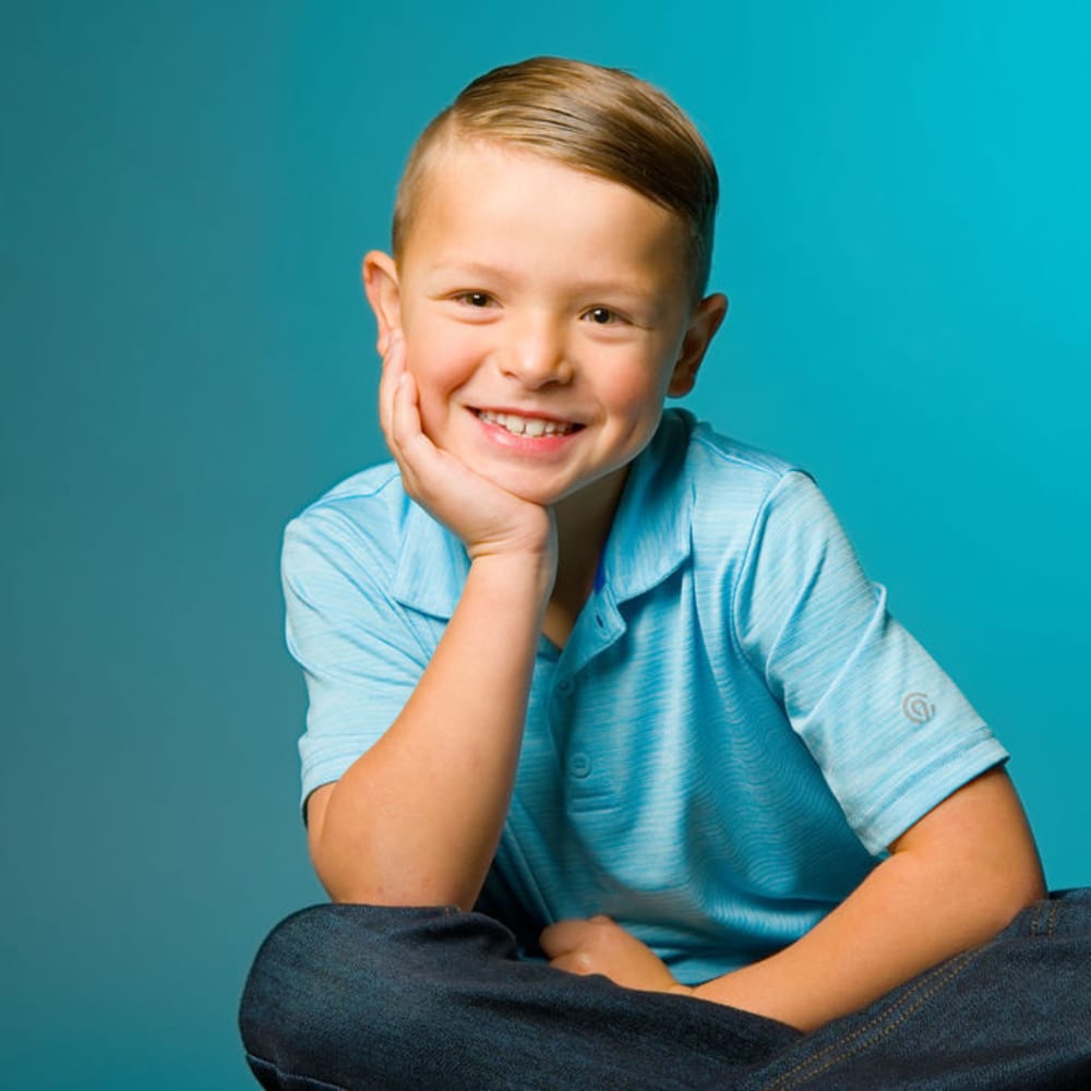 A young boy with a blue collared shirt is smiling with his chin in the palm of his hand.