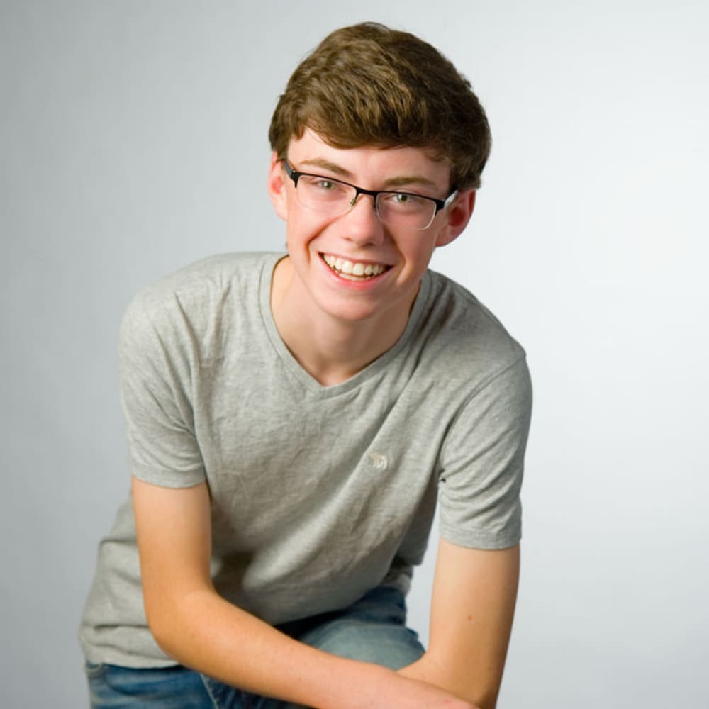 Teenage boy in a gray t-shirt with glasses on is smiling.